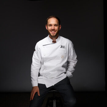 Franz Berlin sitzt lächelnd in einer weißen Kochjacke vor einem dunklen Hintergrund.