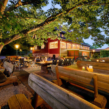 A beer garden with tables and benches in front of a red house at dusk with lights.