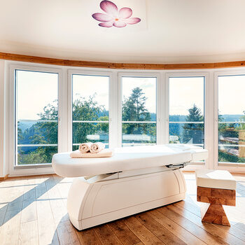 Eine Massageliege steht in einem geräumigen Raum mit großen Panoramafenstern.