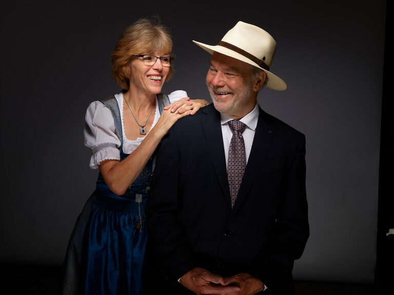Gudrun Berlin in a dirndl and Rolf Berlin in a suit and Panama hat smile at each other against a dark background.