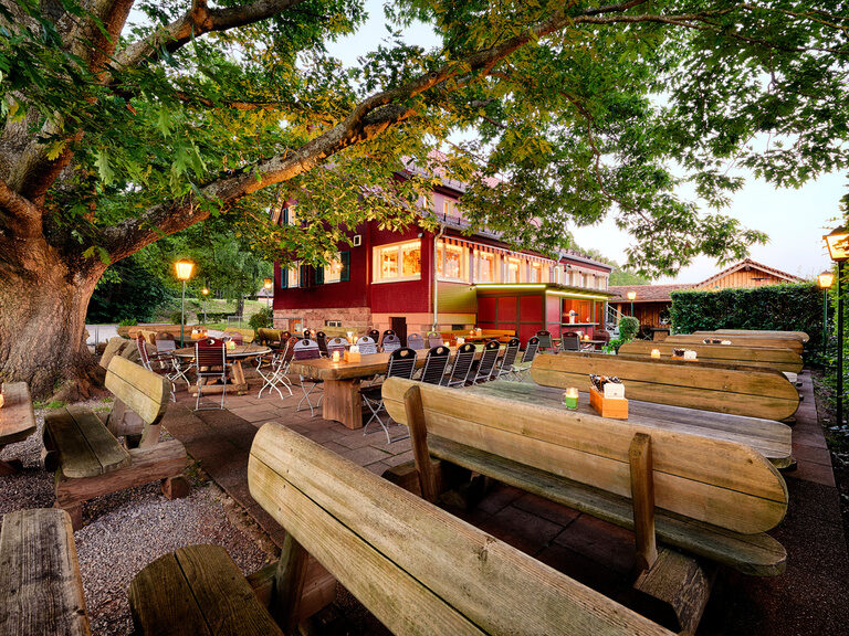 Large beer tables stand in a beer garden in front of a red house.