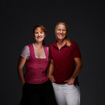 Anja Hügelmann und Kristina Möhrer stehen lächelnd vor einem dunklen Hintergrund.