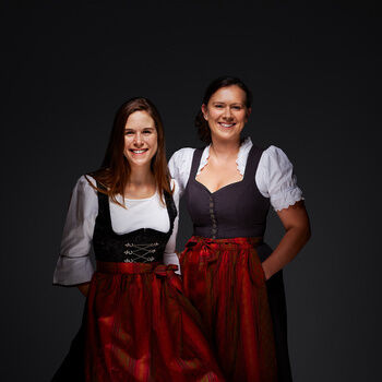 Catrien Müller und Natascha Lötterle, in schwarzen Dirndln mit roten Schürzen, lächelnd vor einem dunklen Hintergrund.