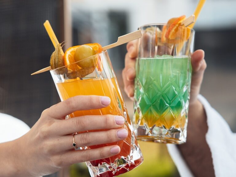 Zwei Hände die bunte Cocktails in den Händen halten und damit anstoßen.