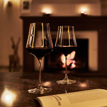 Zwei Gläser Rotwein stehen auf einem Tisch vor einem angefeuerten Kamin.