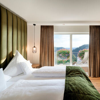 Gemütliches Bett mit grüner Tagesdecke vor einem großen Panoramafenster.