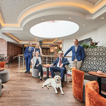 Familie Berlin mit Hund Prinz, stehend und sitzend auf den modernen, orangenen und grauen Sesseln, in der Lounge.
