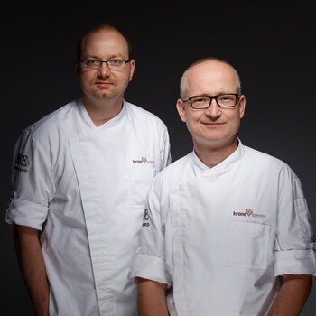 Robby Mittag und Marco Lehnert befinden sich in weißen Kochjacken vor einem dunklen Hintergrund.