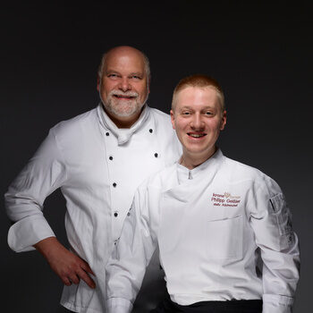Küchenchef Thomas Kling und Stellvertretung Philip Geißler in weißen Kochjacken vor dunklem Hintergrund.