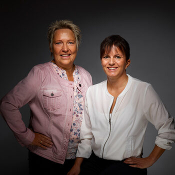 Petra Volle und Anita Nothacker lächelnd vor einem dunklen Hintergrund.