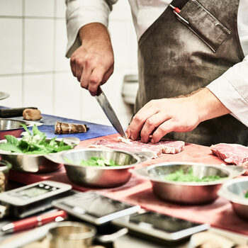 Ein Koch schneidet Fleisch auf einem Brett vor ihm stehen diverse Küchenutensilien sowie verschiedene Kräuter.
