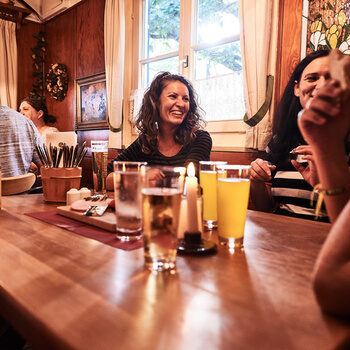 Einige Gäste sitzen um einen Holztisch, auf dem verschiedene Getränkespezialitäten stehen und lachen.