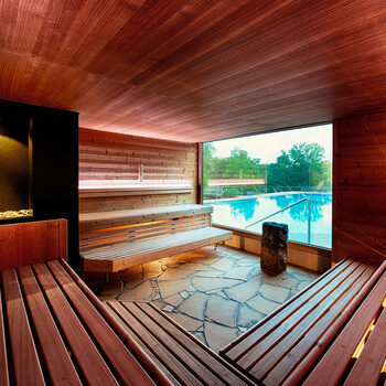 Rotbeleuchtete Sauna mit Sitzgelegenheiten aus Holz und großem Panoramafenster mit Blick auf den Außenpool.