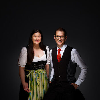 Simona Festa und Oliver Kopp lächeln in Arbeitskleidung vor einem dunklen Hintergrund.