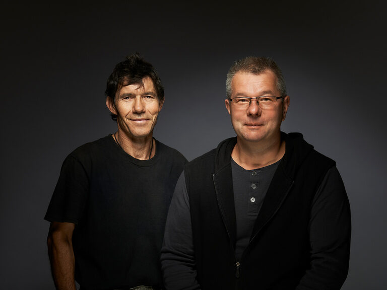 Zwei Männer in handwerklicher Kleidung vor einem dunklen Hintergrund.