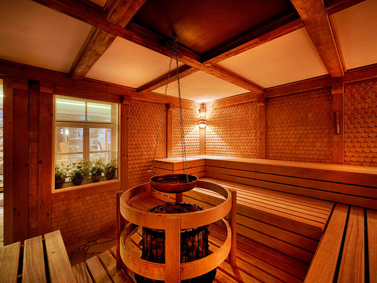 Sitzbänke sowie ein Ofen für Saunaaufgüsse in einer Sauna, deren Wände im Altholzambiente ausgestattet sind.