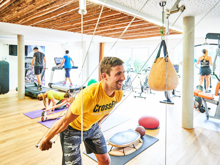 Ein junger Mann sowie weitere Gäste trainieren in einem Fitnessraum an verschiedenen Sportgeräten.