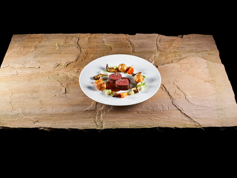 A plate with saddle of venison, almond pralines and seasonal vegetables arranged on a stone slab.