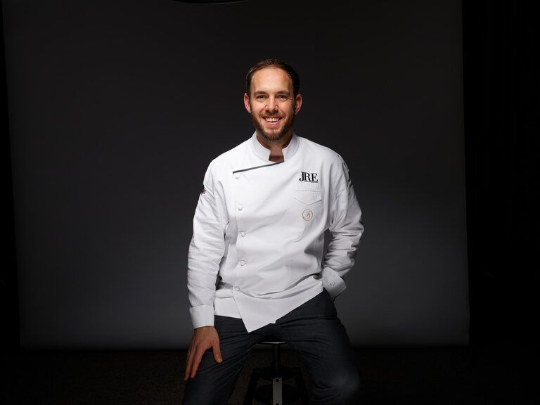 Franz Berlin sits smiling in a white chef's jacket against a dark background.