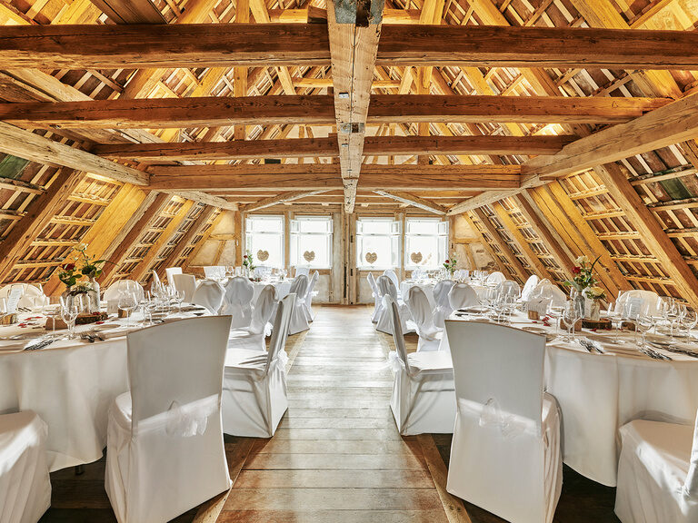 Ein feierlich bestuhlter Dachstuhl mit festlich eingedeckten, runden Tische und, mit weißen Hussen überzogenen, Stühlen.