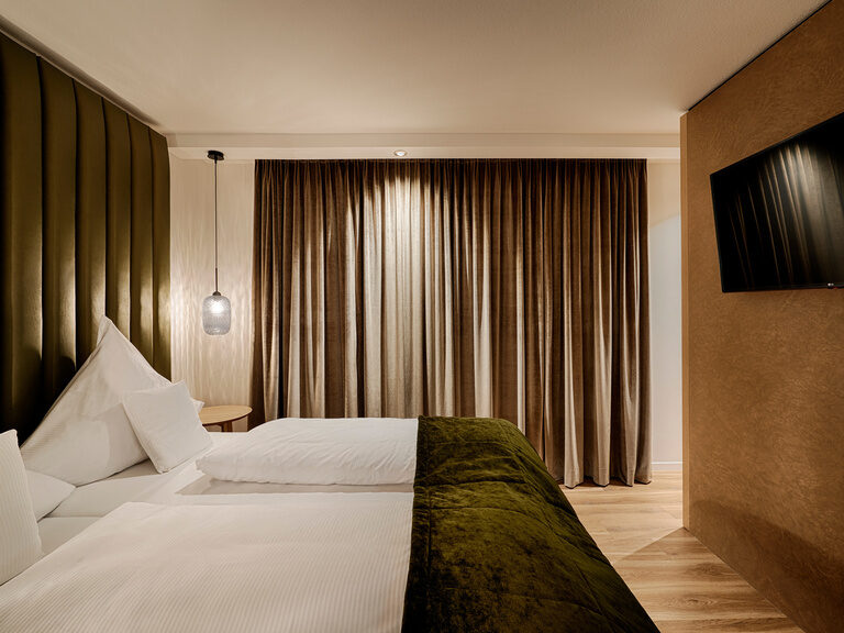 Ein komfortables Bett mit olivgrünen Accessoires steht vor einem Fenster mit geschlossenen Vorhängen.