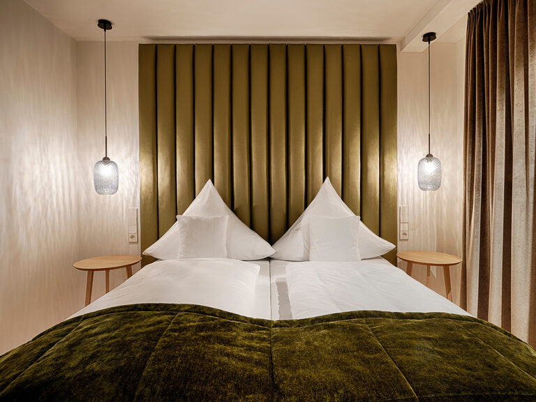 Ein gemütliches Bett mit olivgrünen Accessoires, die für Gemütlichkeit sorgen, steht vor geschlossenen Vorhängen.