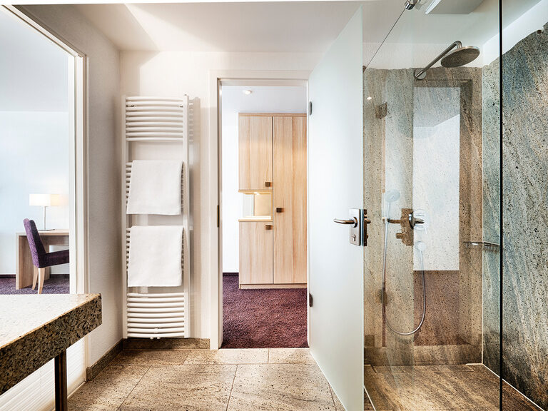 Ein Badezimmer, das durch eine Glasscheibe zum Wohnbereich getrennt ist, mit begehbarer Dusche und Handtuchwärmer.