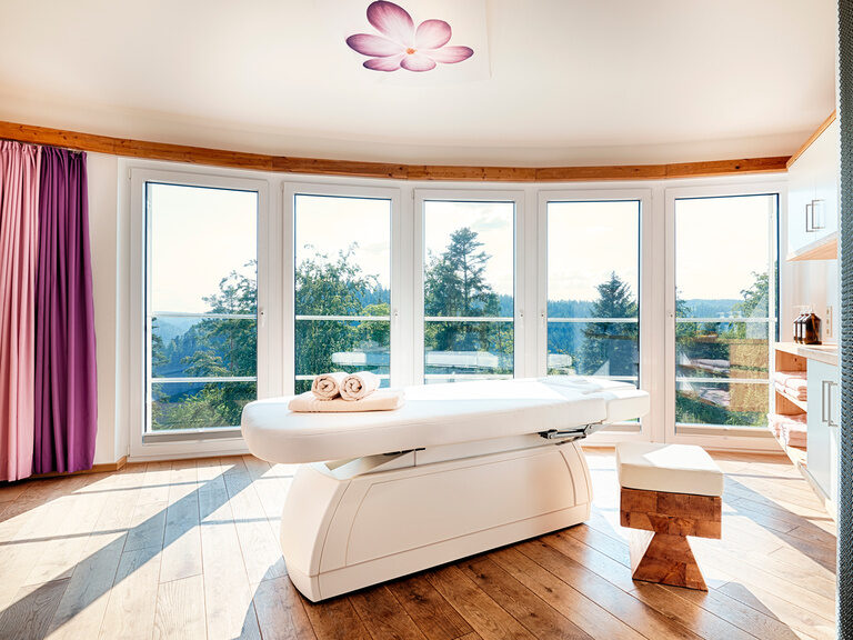 Eine Massageliege steht in einem geräumigen Raum mit großen Panoramafenstern.