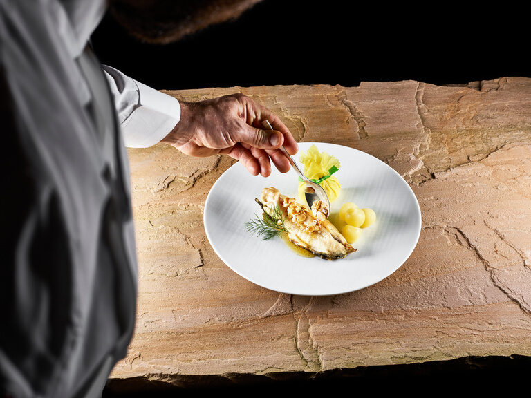 Ein Koch richtet ein Gericht mit Forelle auf einem weißen Teller auf einer Steinplatte an.