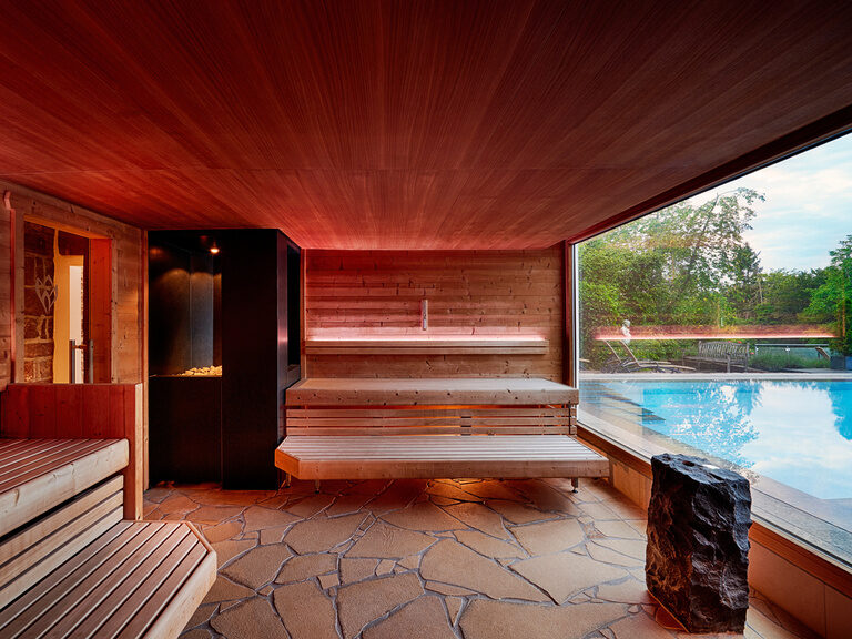 Eine rotbeleuchtete Sauna mit großem Panoramafenster, aus dem der Außenpool des Hotels zu sehen ist.