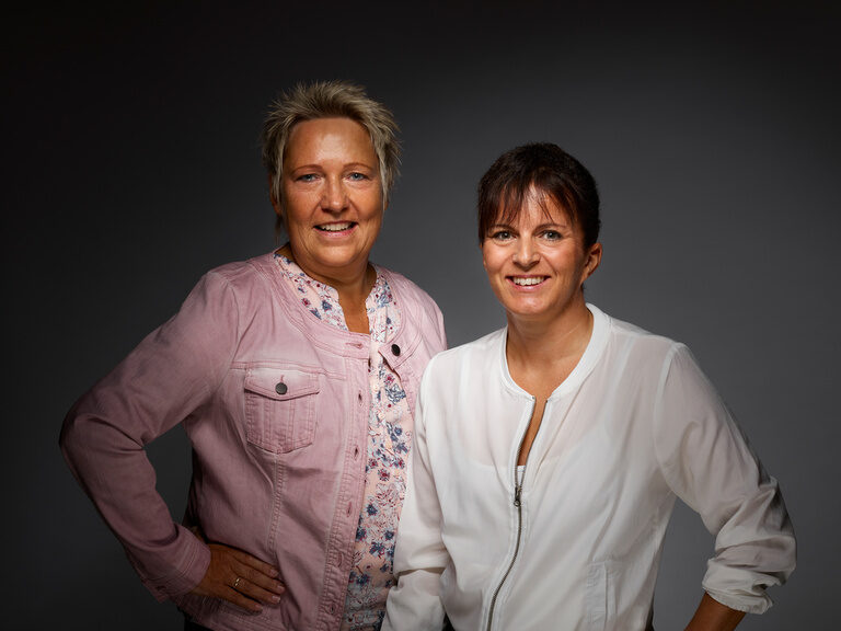 Petra Volle und Anita Nothacker lächelnd vor einem dunklen Hintergrund.
