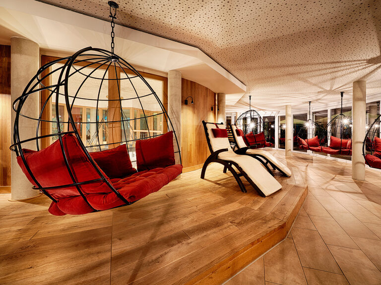 Hanging chairs with red cushions and comfortable wellness loungers in a large room with a continuous window front.