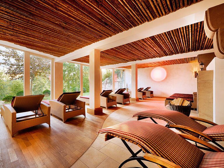 Various loungers in warm colors and a tiled stove in a large relaxation room with a wooden ceiling.