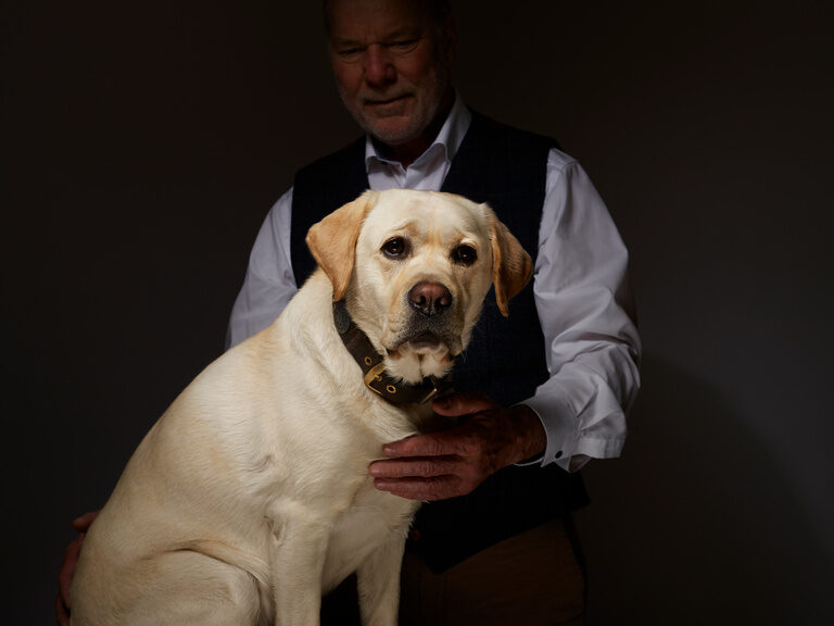 Senior boss, Rolf Berlin, and his white hotel dog Prinz in front of a dark background.