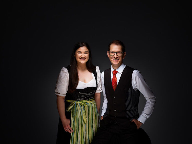 Simona Festa und Oliver Kopp lächeln in Arbeitskleidung vor einem dunklen Hintergrund.