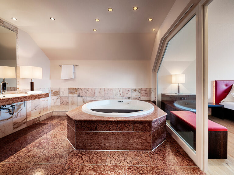 Ein großer Whirlpool in einem Badezimmer, welches durch eine Glasfront zum Schlafbereich getrennt ist.