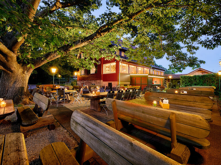 A beer garden with tables and benches in front of a red house at dusk with lights.