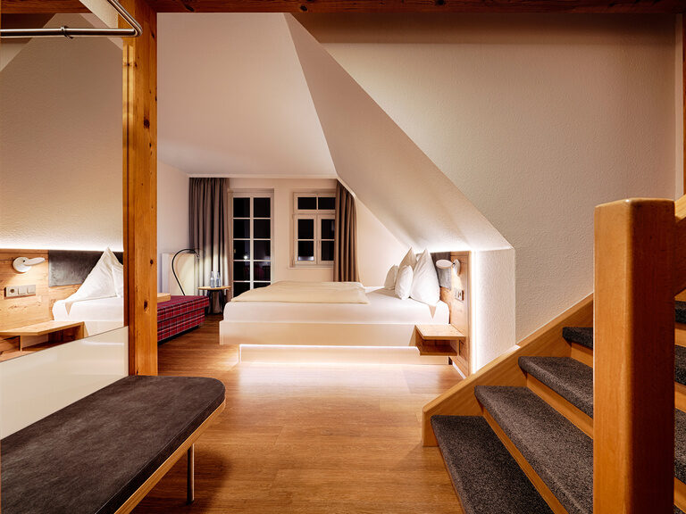 Ein geräumiges, modernes Zimmer in Holzoptik mit Doppelbett, Sitzgelegenheit, Schlafcouch und Treppenaufgang.