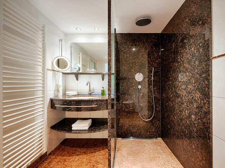 Ein dunkel gehaltenes Badezimmer mit begehbarer Dusche, großem Waschbecken, Spiegeln und Waschutensilien.
