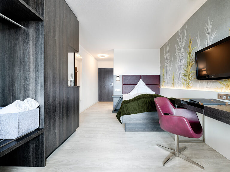 Ein modern eingerichtetes Hotelzimmer mit großem Einzelbett, Kleiderschrank, Schreibtisch und Sessel.