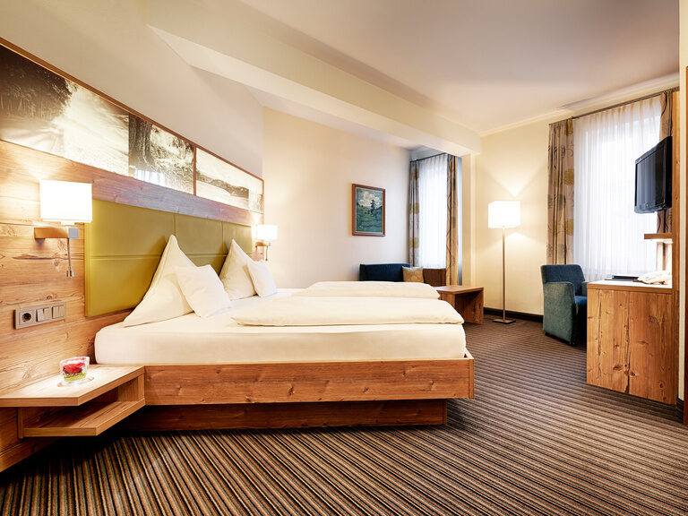 Im hellen Landhausstil eingerichtetes Hotelzimmer mit Doppelbett, Teppichboden, Sitzgelegenheit sowie Schreibtisch.