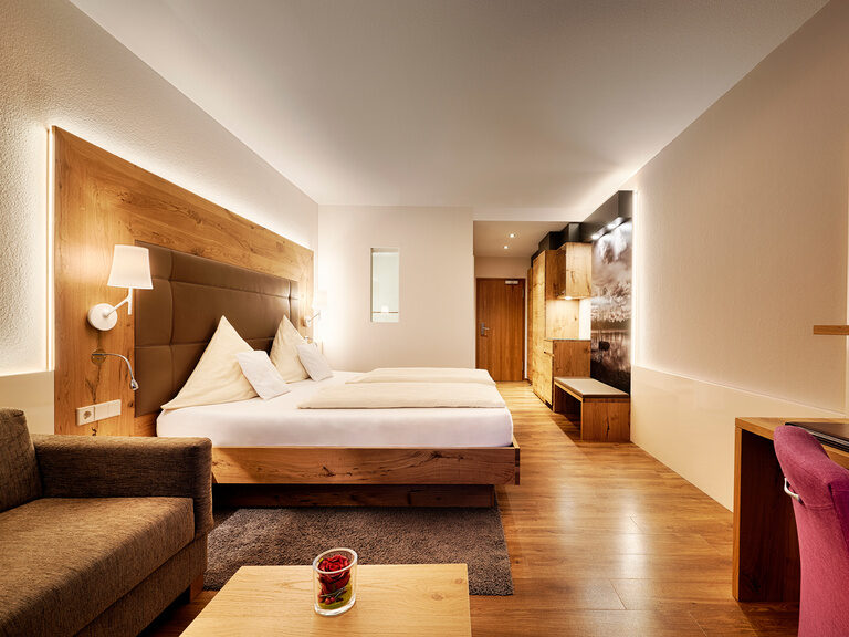 Ein Doppelbett, Möbeln in hellem Holz einer gemütlichen Couch sowie Schreibtisch in gemütlich beleuchteter Atmosphäre.