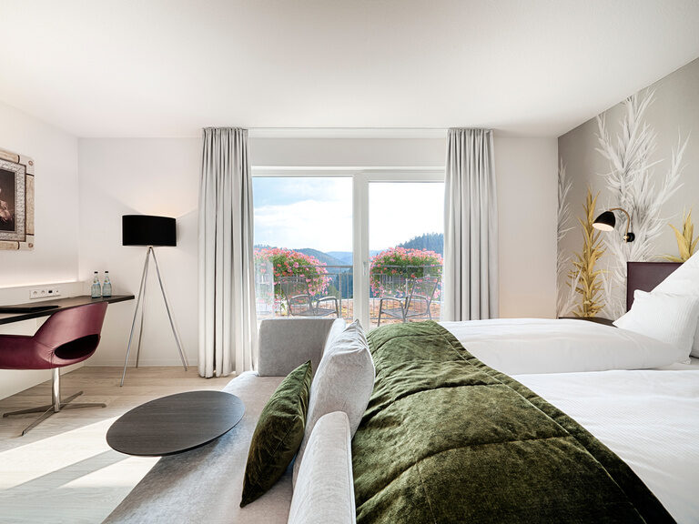 Ein großzügiges Bett, auf dem weiße Bettwäsche und eine grüne Tagesdecke liegt in einem lichtdurchfluteten Hotelzimmer.